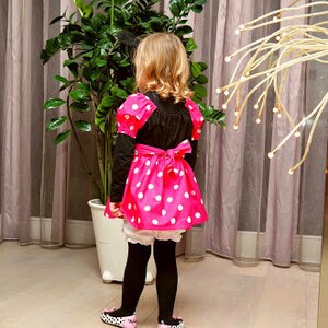 Op de afbeelding: Een jong persoon draagt een roze en wit gebloemde jurk met een zwart bovenstuk en een wit onderrok. De jurk heeft een strik in de taille. De persoon draagt zwarte panty's en zwarte schoenen.