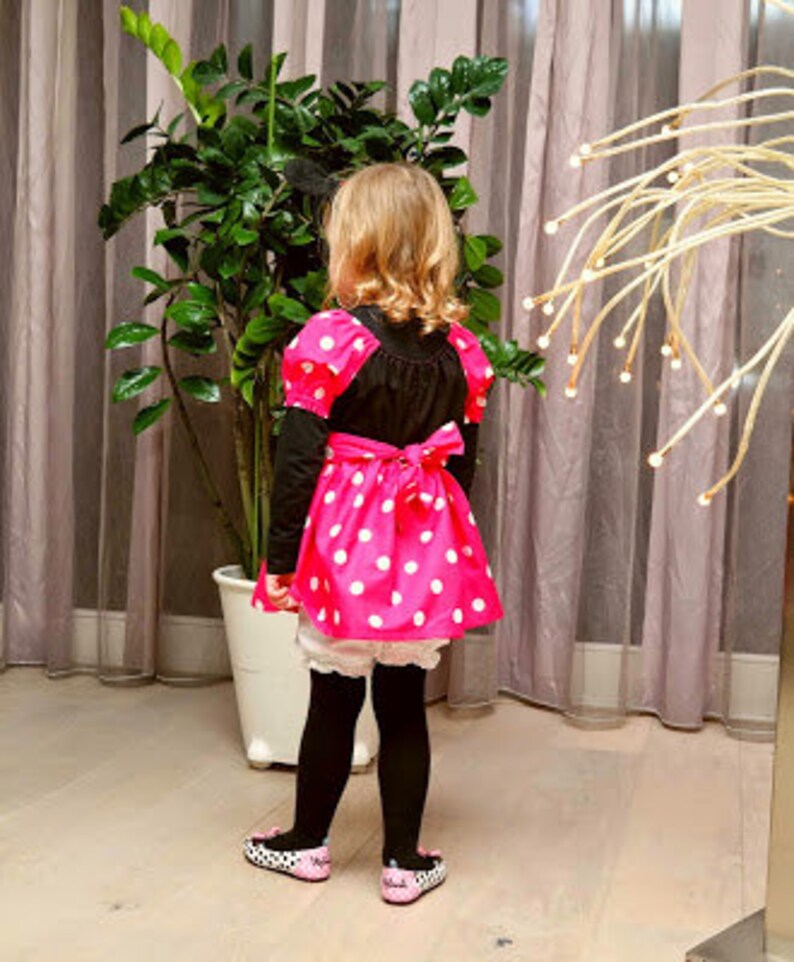 Op de afbeelding: Een jong persoon draagt een roze en wit gebloemde jurk met een zwart bovenstuk en een wit onderrok. De jurk heeft een strik in de taille. De persoon draagt zwarte panty's en zwarte schoenen.