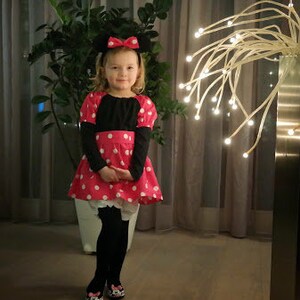 Op de afbeelding: Een jong meisje verkleed als Minnie Mouse, met een rood-wit geblokt jurkje met zwarte mouwen en een zwart strik-hoofdband. Ze staat voor een witte muur met een decoratieve lichtinstallatie achter haar.