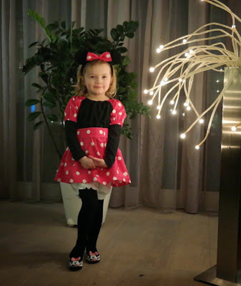 Op de afbeelding: Een jong meisje verkleed als Minnie Mouse, met een rood-wit geblokt jurkje met zwarte mouwen en een zwart strik-hoofdband. Ze staat voor een witte muur met een decoratieve lichtinstallatie achter haar.