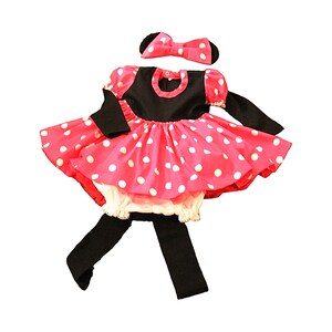 Op de afbeelding: Een roze en witte stippenjurk met zwarte mouwen en een zwart hoofdband met een strik. De jurk heeft een witte onderrok.