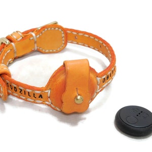 May include: Orange leather dog collar with white stitching and the word "GODZILLA" embossed on the strap. The collar has a small, round, orange leather pouch with a gold button. A black round device with a white button is also pictured.