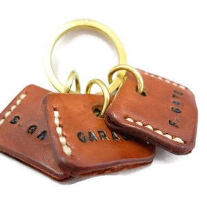 May include: A set of three brown leather keychains with white stitching and brass rings. Each keychain has a square shape and features black stamped text: "S. GA", "GARAGE", and "F. GATE".