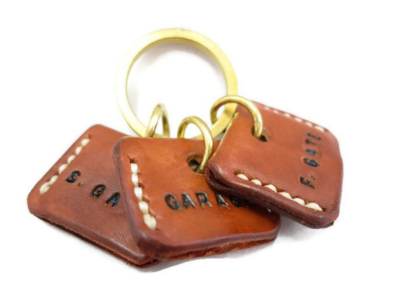 May include: A set of three brown leather keychains with white stitching and brass rings. Each keychain has a square shape and features black stamped text: "S. GA", "GARAGE", and "F. GATE".