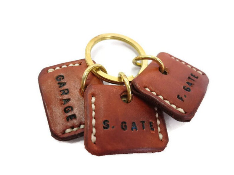 May include: A set of three brown leather keychains with brass rings. Each keychain has a square shape with white stitching around the edges. The keychains are stamped with the words "GARAGE", "S. GATE", and "F. GATE" in black.