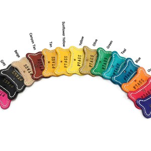 May include: A collection of colorful leather pet tags in various shades, including pink, black, grey, beige, tan, yellow, green, blue, orange, red, and violet. Each tag is stitched with white thread and features the word "PTAGS".