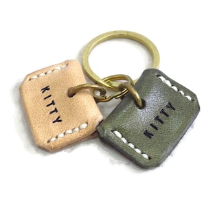 Peut inclure: Deux porte-clés en cuir avec le mot "KITTY" gravé sur chacun. Un porte-clés est beige avec des coutures blanches et l'autre est vert avec des coutures blanches.