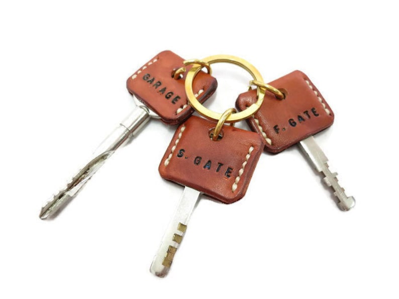 May include: Three keys with brown leather key fobs attached to a gold-colored ring. Each fob is square with white stitching and stamped with the words "GARAGE", "F. GATE", and "S. GATE". The keys have silver metal blades.
