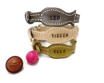 May include: Three leather dog collars with the names "Leo", "Tigger", and "Elle" embroidered on them. The collars are stacked on top of each other, with the top collar being brown, the middle collar being tan, and the bottom collar being olive green. There is a pink bell attached to the bottom collar.