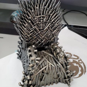 Könnte beinhalten: Eine silberfarbene Miniatur-Replik des Eisernen Throns aus der Fernsehserie Game of Thrones. Der Thron besteht aus Schwertern und ist ein beliebtes Sammlerstück für Fans der Serie.