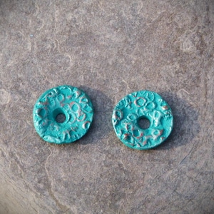 Pode incluir: Duas contas de cerâmica de cor turquesa azul em forma de rosquinha com um design texturizado em relevo. As contas têm um furo no centro.