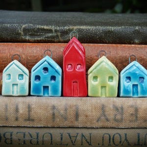 May include: A collection of five ceramic house ornaments in various colors. The houses are in shades of blue, green, and red, with small windows and doors. Each ornament has a metal loop for hanging, perfect for holiday decor.