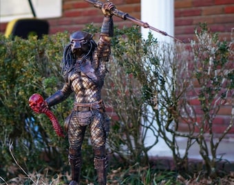 Op de afbeelding: Een Predator-actiefiguur dat een speer en een schedel vasthoudt. De figuur draagt een camouflagepak en heeft een rood-zwarte schedel in zijn hand. De figuur staat op een voetstuk met het woord "PREDATOR" erop.