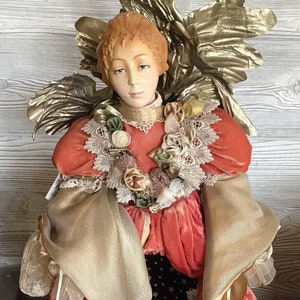 Può includere: Una statuetta di angelo in porcellana con ali in tessuto di velluto rosso e oro. L'angelo indossa un abito in velluto rosso con bordi dorati e pizzo. L'angelo è seduto su un cuscino in velluto verde con un bordo dorato.