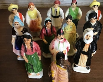 Puede incluir: Una colección de 13 figuritas de cerámica que representan personas de diferentes culturas con ropa tradicional. Las figuritas están pintadas en varios colores, incluyendo rojo, azul, verde, amarillo y marrón. Están de pie sobre una superficie de madera.