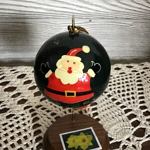 Peut inclure: Une boule de Noël ronde et verte avec une image peinte du Père Noël. La boule est sur un support en bois avec une image de jonquilles jaunes.