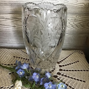 Peut inclure: Un vase en verre taillé transparent avec un motif floral en forme de cœur. Le vase a un bord festonné et est décoré de feuilles et de fleurs gravées. Une branche de fleurs bleues et blanches repose devant le vase.