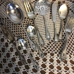 以下が含まれることがあります： スプーン、フォーク、穴あきスプーン、バターナイフなど、アンティークの銀製サービング用具の詰め合わせ。 銀食器はハンドルに華やかな花のディテールがあり、白いレースのテーブルクロスの上に配置されています。