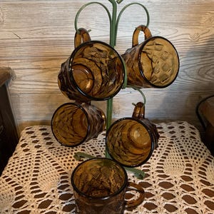 May include: A set of five amber-coloured glass mugs with a textured diamond pattern, hanging on a green metal stand. Each mug features a sturdy handle and a faceted exterior design, with four mugs suspended from the hooks and one resting at the base.