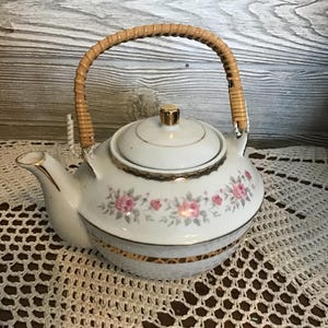 Puede incluir: Tetera de porcelana blanca con asa trenzada y detalles dorados. La tetera presenta un estampado floral de rosas rosas y hojas verdes. La tapa está rematada con un pomo dorado. La tetera está sobre un tapete de encaje blanco.