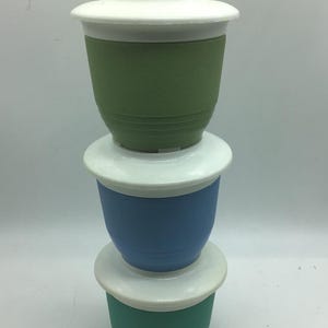 May include: Three stackable plastic containers with white lids. The top container is green, the middle container is blue, and the bottom container is a darker green.
