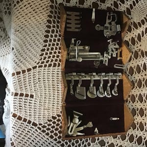 May include: A wooden sewing machine attachment case with a dark purple velvet interior lining. The case contains various silver-colored metal presser feet, hemmers, and specialized sewing components organized in rows within the velvet-lined compartments.