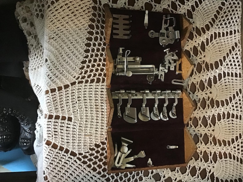 May include: A wooden sewing machine attachment case with a dark purple velvet interior lining. The case contains various silver-colored metal presser feet, hemmers, and specialized sewing components organized in rows within the velvet-lined compartments.