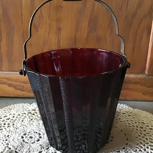 May include: A dark red glass ice bucket with a metal handle. The bucket has a fluted design and is sitting on a white crocheted doily.