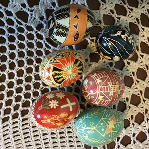 May include: Six intricately decorated Easter eggs in a variety of colours. The eggs feature detailed patterns and designs, including orange, red, green, and blue. They are displayed on a white, open-weave surface.