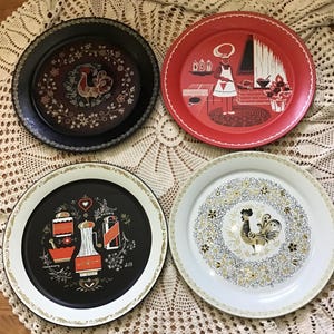 May include: Four round metal serving trays. One black tray features a rooster and floral border. One red tray depicts a chef in a kitchen. One black tray displays kitchen spice jars and herbs labelled parsley, rosemary, and dill. One white tray shows a rooster.