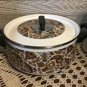 Op de afbeelding: Een witte emaille pan met een bruin en gouden paisley-patroon. De pan heeft een deksel met een zilveren rand en een zwarte knop.