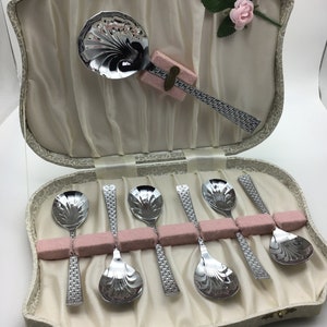 Puede incluir: Un juego de seis cucharaditas de plata con mangos adornados en una caja blanca y dorada con forro rosa. La caja tiene una rosa en la parte superior.