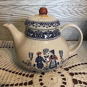 Puede incluir: Tetera de cerámica color crema con un diseño floral azul y blanco y un pomo rojo en la tapa. La tetera tiene un asa y un pico, y un borde decorativo. El cuerpo de la tetera tiene una escena pintada de dos figuras.