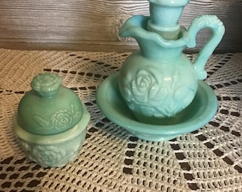 May include: A vintage, light blue glass vanity set featuring a small pitcher with a rose design, a matching bowl, and a lidded jar. The set is displayed on a crocheted doily, showcasing a classic, antique aesthetic.