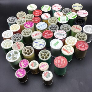 Puede incluir: Una colección de carretes de hilo de varios colores, incluyendo verde, rojo, amarillo y marrón. Los carretes están etiquetados con marcas como Coats, Esprit y Drima. Algunos carretes tienen una etiqueta blanca con texto negro, mientras que otros tienen una etiqueta roja con texto blanco. Los carretes están dispuestos en un patrón aleatorio sobre una superficie negra.