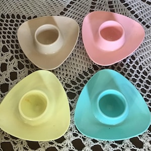May include: Four pastel-coloured egg cups, each with a triangular base and a central circular recess. The colours are beige, pink, yellow, and teal. The egg cups are displayed on a white, crocheted surface. Each cup is designed to hold an egg.
