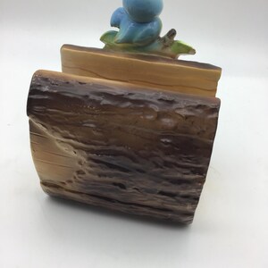 May include: A ceramic log holder with a blue bird perched on top. The log holder is painted to resemble a tree trunk with brown and tan bark.