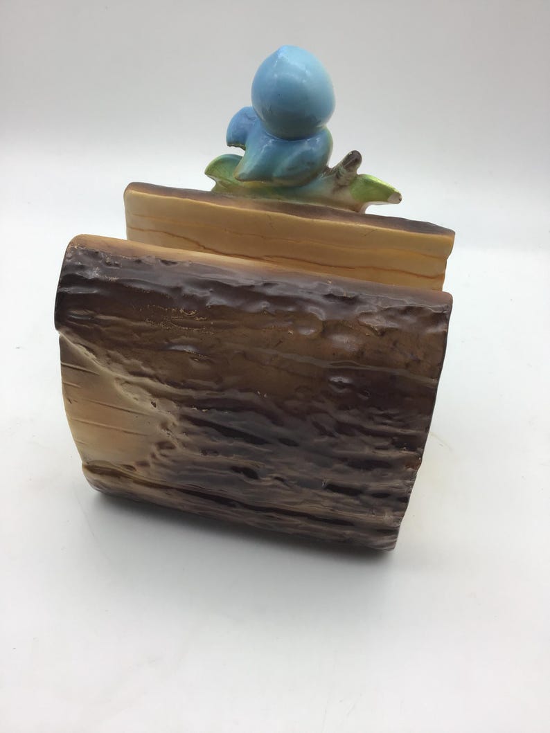 May include: A ceramic log holder with a blue bird perched on top. The log holder is painted to resemble a tree trunk with brown and tan bark.