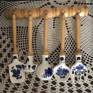 Könnte beinhalten: Ein Set aus fünf Küchenutensilien mit Holzgriffen und weißen Keramikköpfen. Die Keramikköpfe sind mit blauen Blumenmustern verziert. Die Utensilien hängen an einer Holzleiste mit Metallringen.