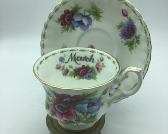 May include: A white teacup and saucer with a floral design and gold trim. The teacup has the word "March" written on it. The saucer has a floral design with a blue and purple flower.