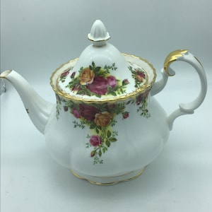 Peut inclure: Une théière en porcelaine blanche avec une bordure dorée et un motif floral composé de roses rouges, jaunes et roses. La théière a un couvercle avec un bouton sur le dessus et une poignée sur le côté.