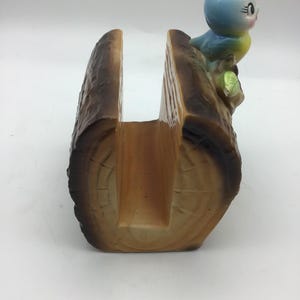 May include: A ceramic napkin holder shaped like a tree log with a blue bird perched on top. The bird has yellow accents and a green leaf.