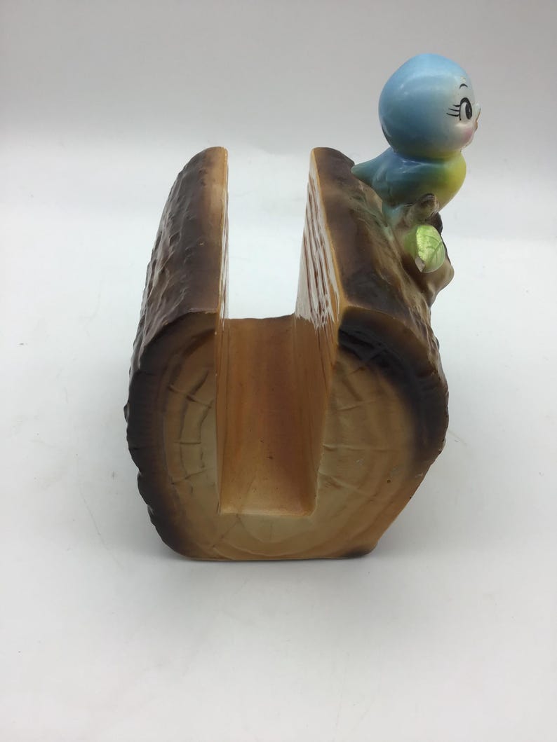 May include: A ceramic napkin holder shaped like a tree log with a blue bird perched on top. The bird has yellow accents and a green leaf.