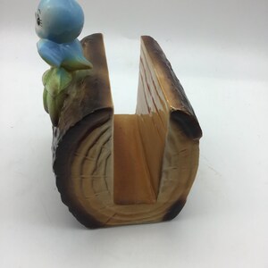 May include: A ceramic napkin holder shaped like a log with a blue bird perched on top. The log is brown with a wood grain pattern and has a slot for napkins.
