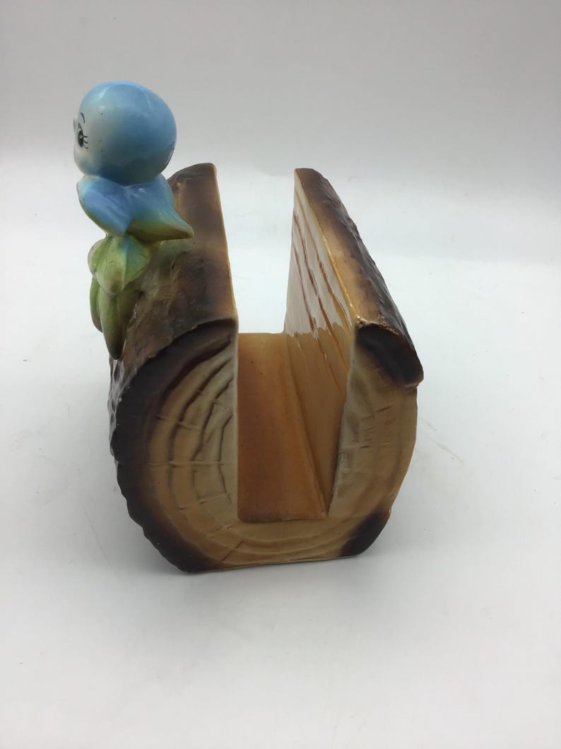May include: A ceramic napkin holder shaped like a log with a blue bird perched on top. The log is brown with a wood grain pattern and has a slot for napkins.