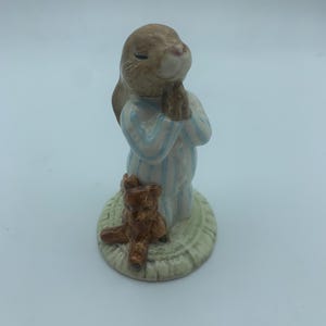 May include: A ceramic figurine of a brown rabbit wearing blue and white striped pyjamas, with its paws clasped together in prayer. The rabbit is standing on a green and white base with a brown teddy bear sitting at its feet.