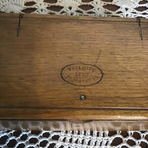 May include: A rectangular wooden box with a visible wood grain texture. An oval stamp on the lid features the text PATENTED 1935 FEBRUARY 19. The box has a small metal pin centered below the stamp and two vertical lines etched into the top edge.