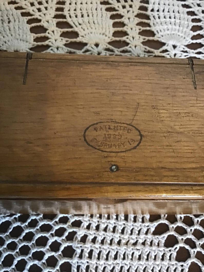 May include: A rectangular wooden box with a visible wood grain texture. An oval stamp on the lid features the text PATENTED 1935 FEBRUARY 19. The box has a small metal pin centered below the stamp and two vertical lines etched into the top edge.