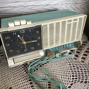 May include: A vintage turquoise and white clock radio with a black clock face and a dial for tuning radio stations. The radio has a cord and is sitting on a white crocheted doily.