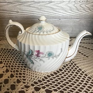 May include: A white porcelain teapot with a spiral design and floral patterns in pink, green, and blue. The teapot has a curved handle and spout, with gold trim. The lid is topped with a small finial.